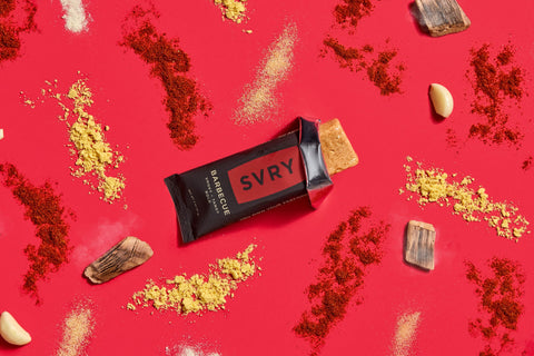 SVRY barbecue protein bar surrounded by spices, garlic cloves and smoked wood chips on a red background