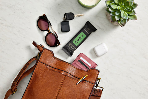 Brown leather handbag with sunglasses, keys, SVRY bar, and white earbuds on a light surface.