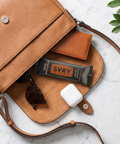 Brown leather bag with sunglasses, an everything bagel protein bar, Airpods and a wallet spilling out onto a marble counter
