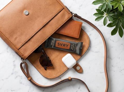 Brown leather bag with sunglasses, an everything bagel protein bar, Airpods and a wallet spilling out onto a marble counter