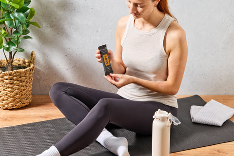 Woman sitting on a yoga mat holding a SVRY coconut curry protein bar in a home setting.