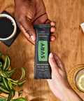 Person holding an SVRY bar on a wooden table with coffee, glass of water, and plant.