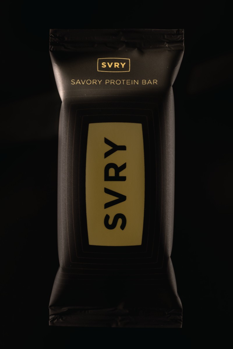 An unlabeled black SVRY savory protein bar wrapper standing against a black background, with a soft diagonal sliver of light falling across the top of the pack where the next flavor name will be printed.
